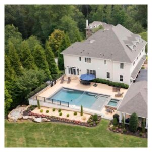 Overhead view of a custom swimming pool behind a home. The edge of it is trimmed with a copse of trees on a hill.